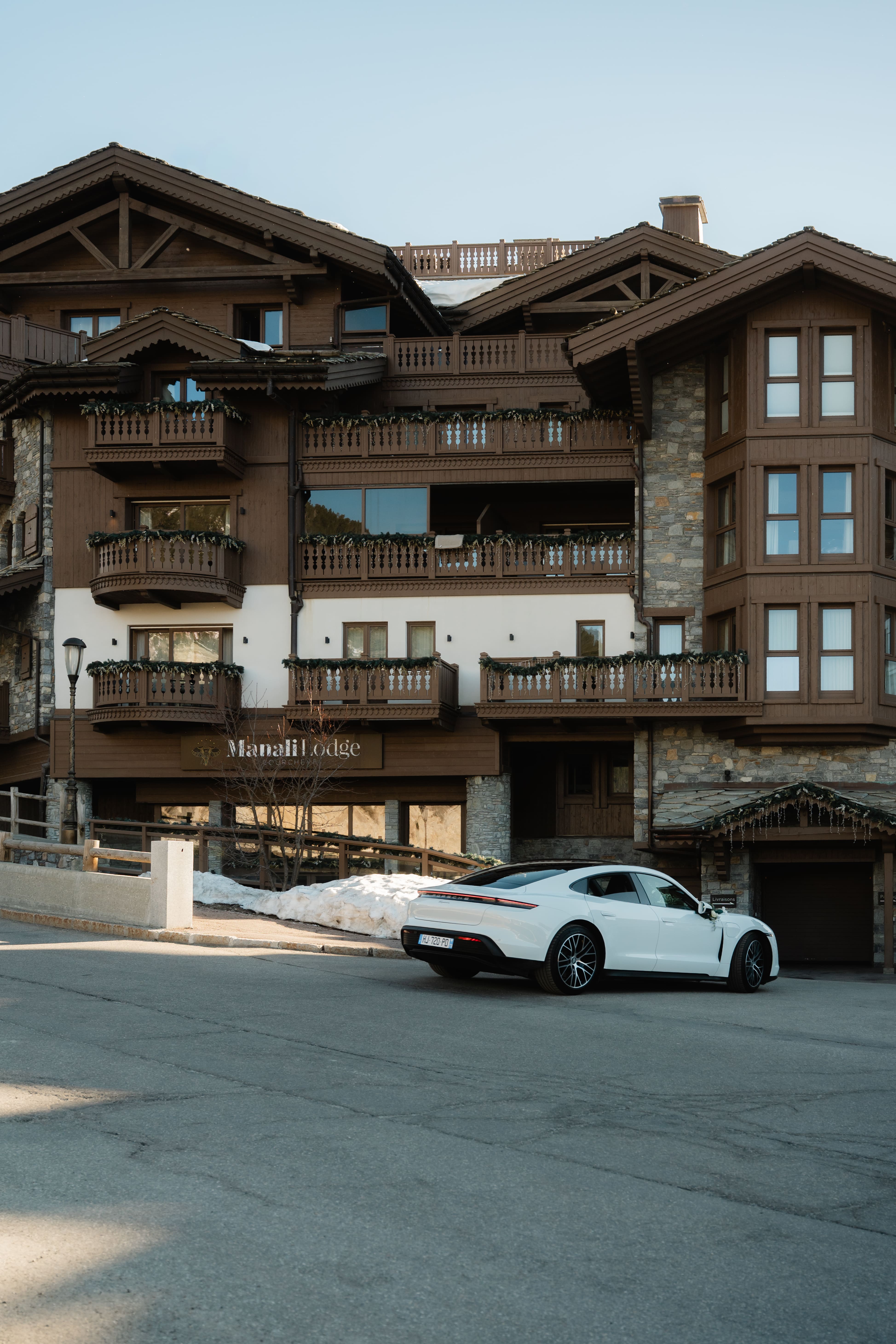 Manali Lodge, chalet en bois sombre de Courchevel 1850, Porsche Taycan blanche garée devant, lumière dorée de fin d'après-midi.