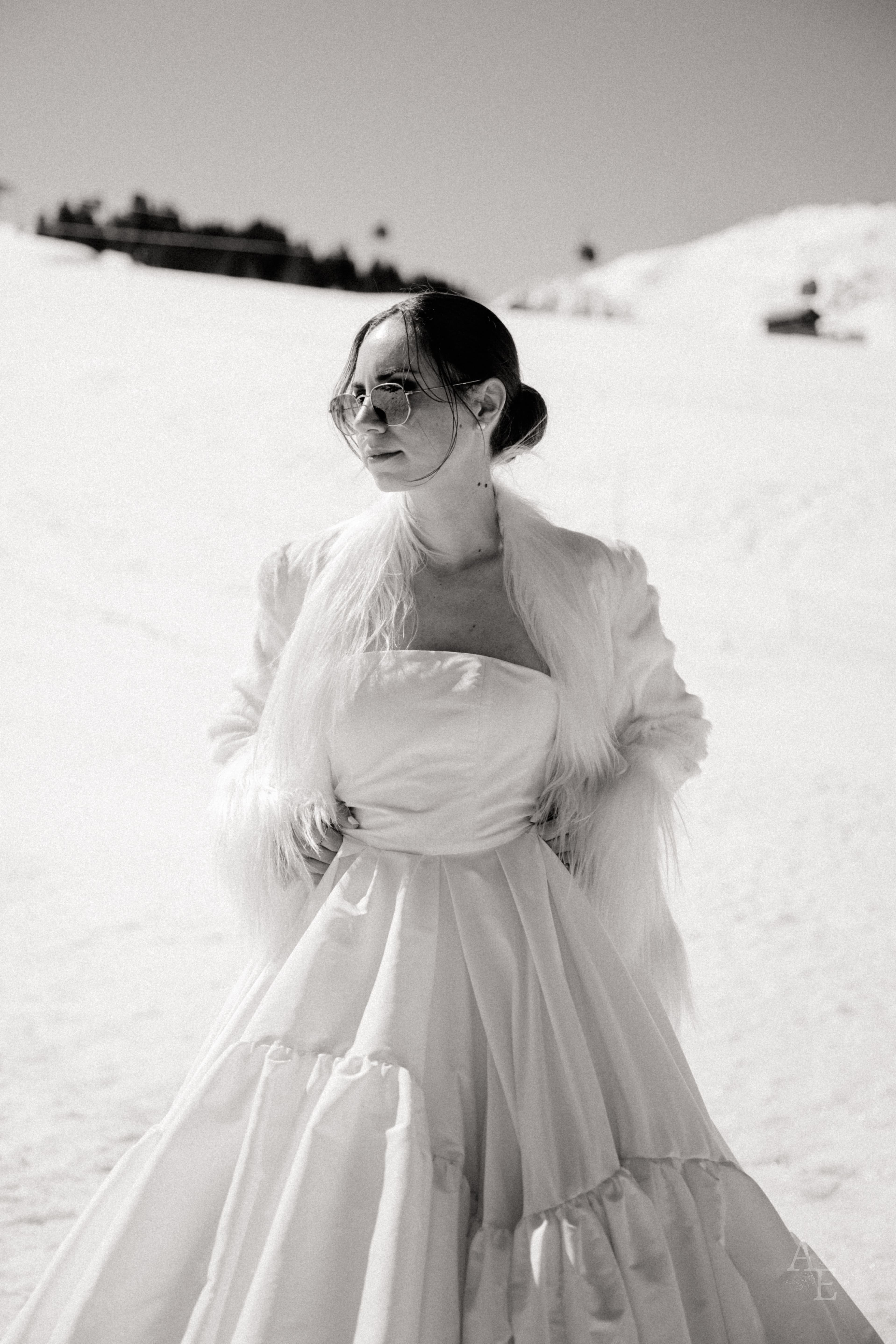 Portrait noir et blanc d'une mariée en robe blanche, étole de plumes et lunettes rondes, dans la neige des 3 Vallées.