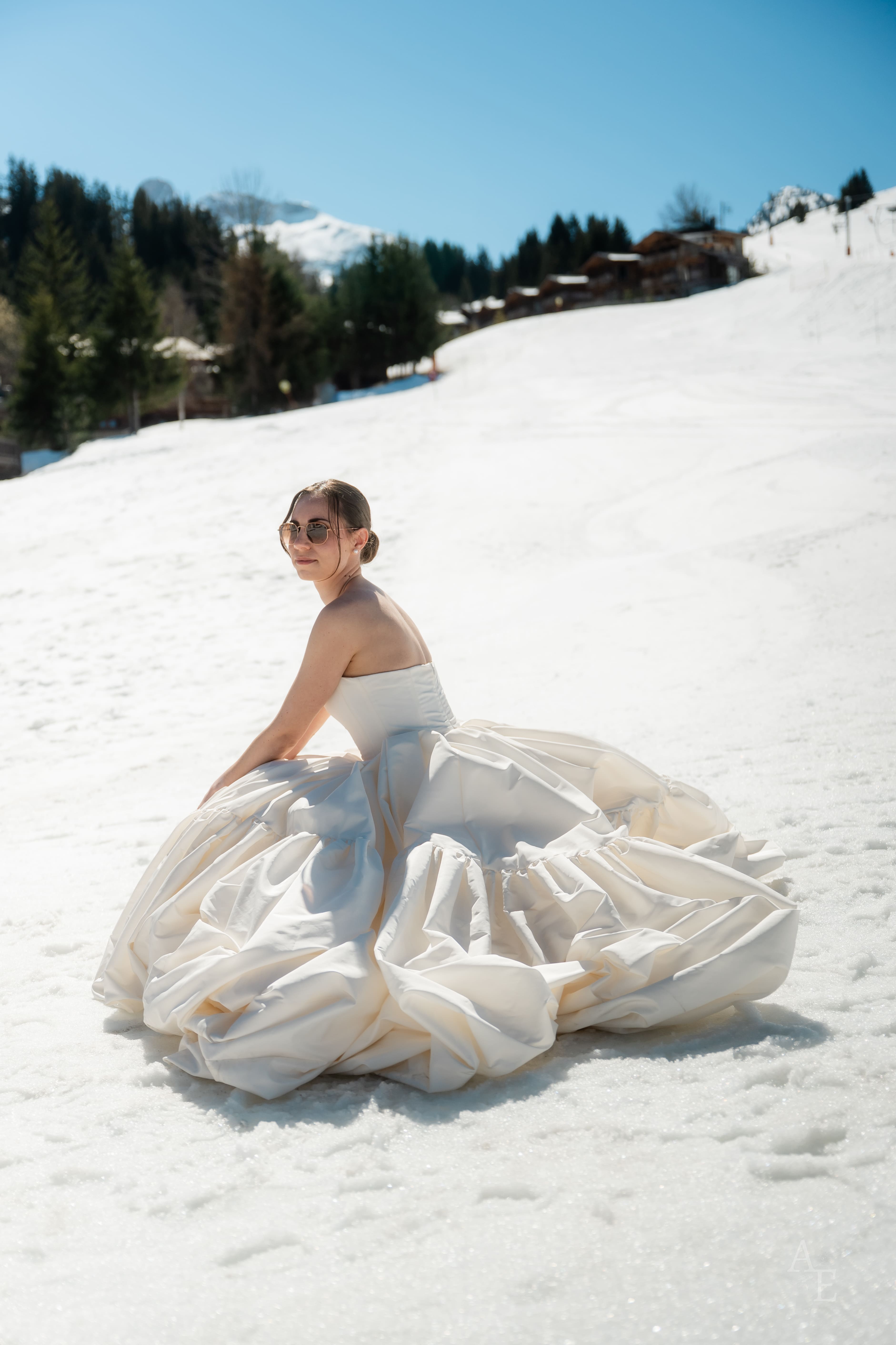 Mariée en robe couture forme meringue, assise sur la neige d'une piste de Courchevel, plein soleil.